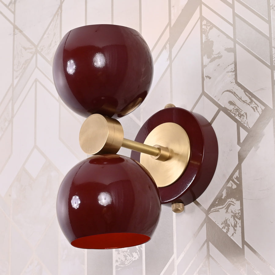 Brass Leaf Studio double globe wall sconce in deep burgundy with brushed brass accents, mounted on geometric patterned wall.