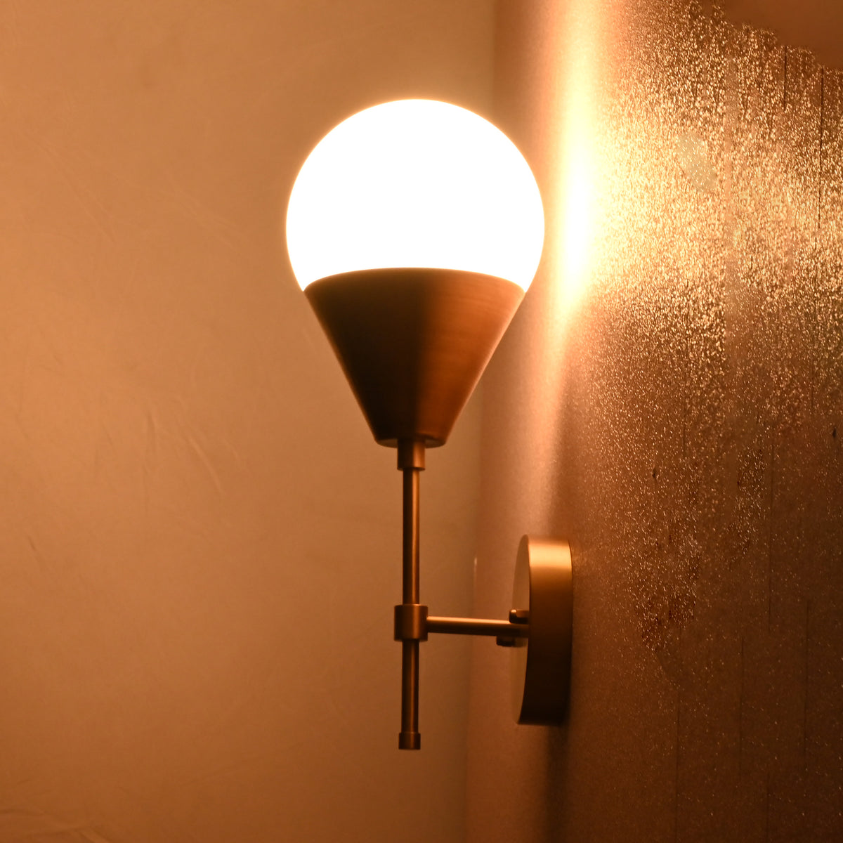 Brass wall sconce glowing warmly in dim light, featuring frosted globe shade and brass cone holder.