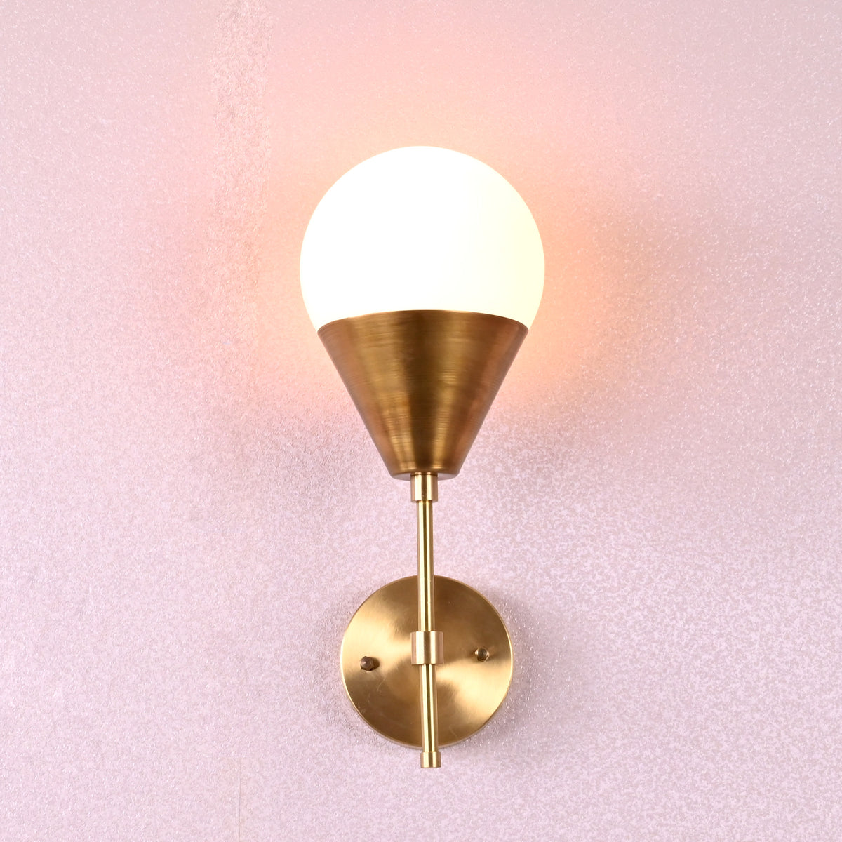 Brass wall lamp with cone base and frosted globe shade in brushed finish, mounted on pink textured wall.