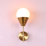 Brass wall lamp with cone base and frosted globe shade in brushed finish, mounted on pink textured wall.