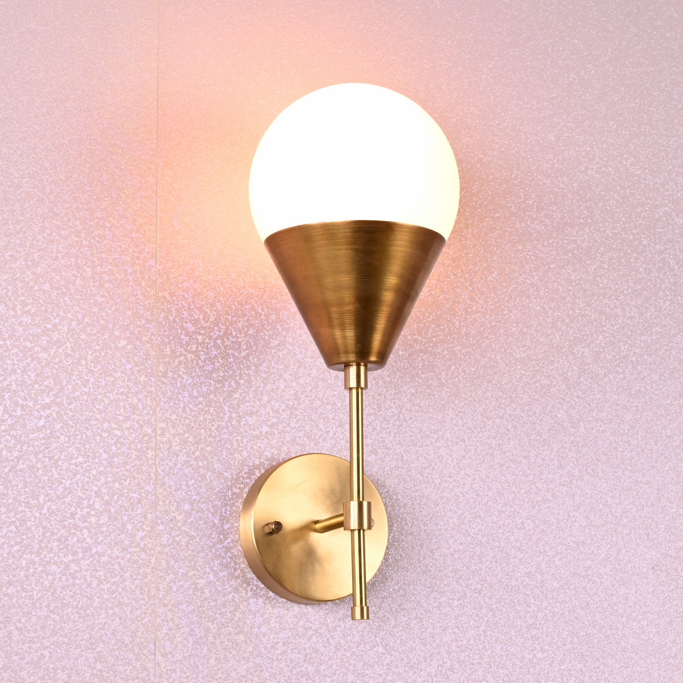 Modern brass wall sconce with globe shade and cone base in brushed brass finish, mounted on textured wall.