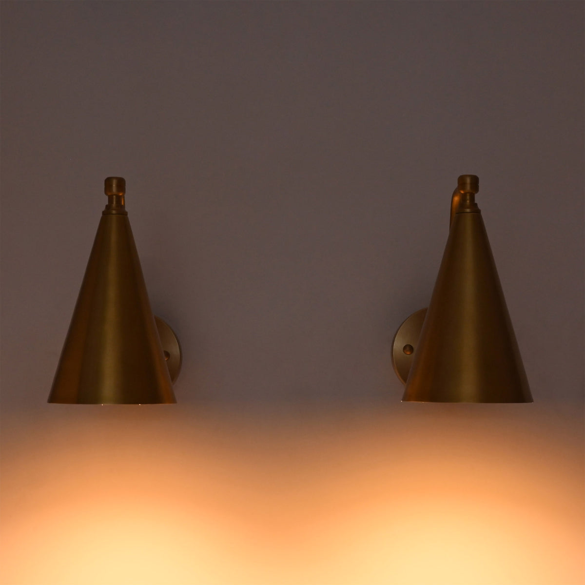 A pair of modern cone-shaped wall sconces in a brushed brass finish, mounted on a wall and casting a warm orange glow downwards.