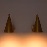 A pair of modern cone-shaped wall sconces in a brushed brass finish, mounted on a wall and casting a warm orange glow downwards.