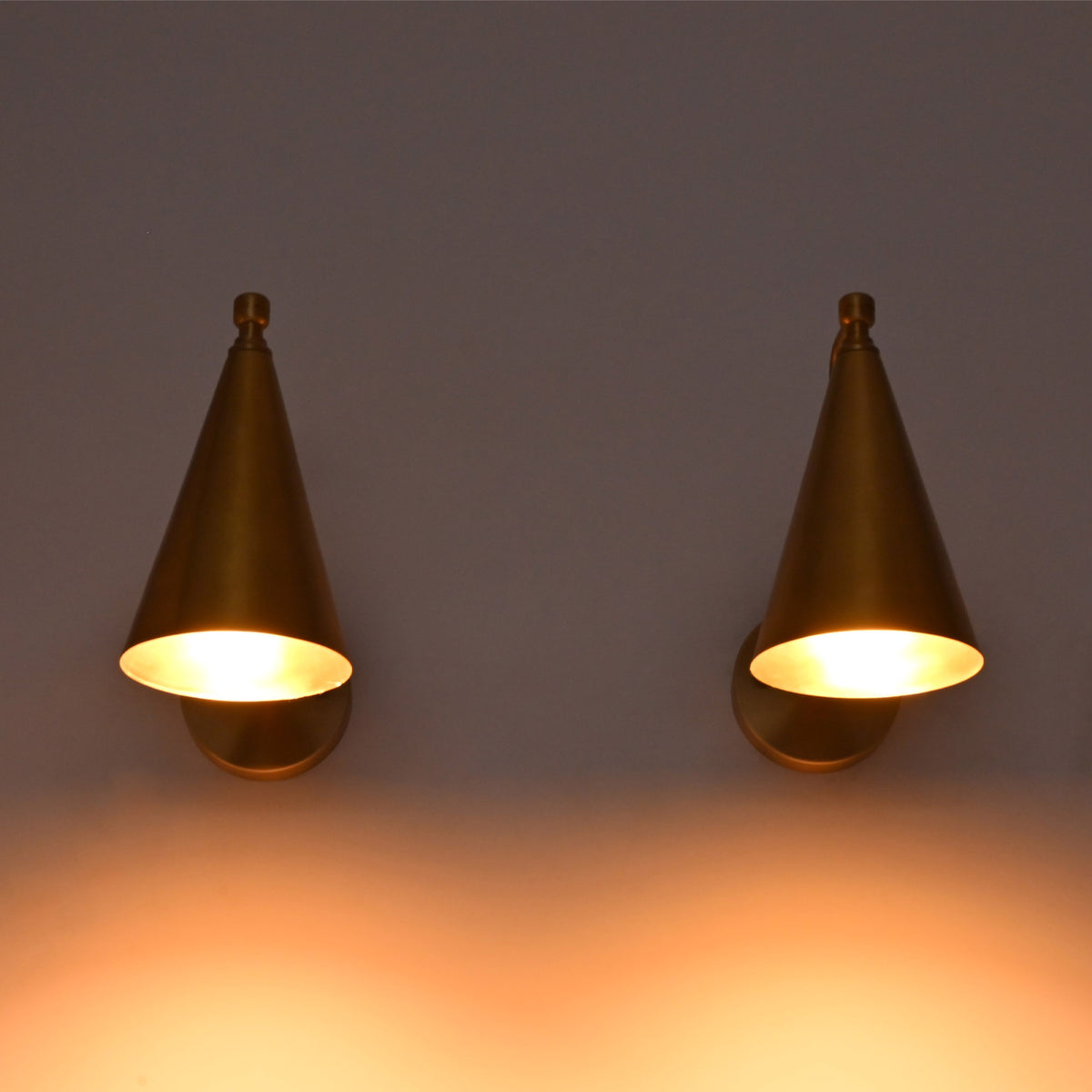 Two matching cone-shaped wall sconces in a warm brass or bronze finish, mounted side by side on a wall and casting a soft warm glow downward.