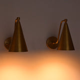 Two brass wall sconces with cone-shaped shades and adjustable curved arms, mounted on a wall and casting a warm orange light downward.