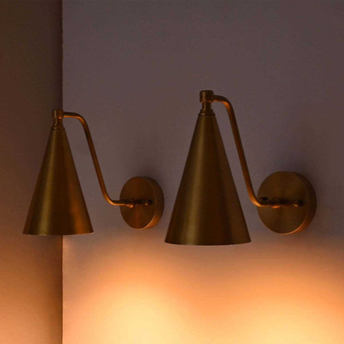Two brass wall sconces with cone-shaped shades and adjustable curved arms, mounted on a wall and casting a warm orange glow.