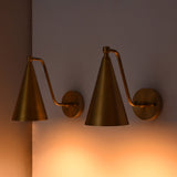 Two brass wall sconces with cone-shaped shades and adjustable curved arms, mounted on a wall and casting a warm orange glow.
