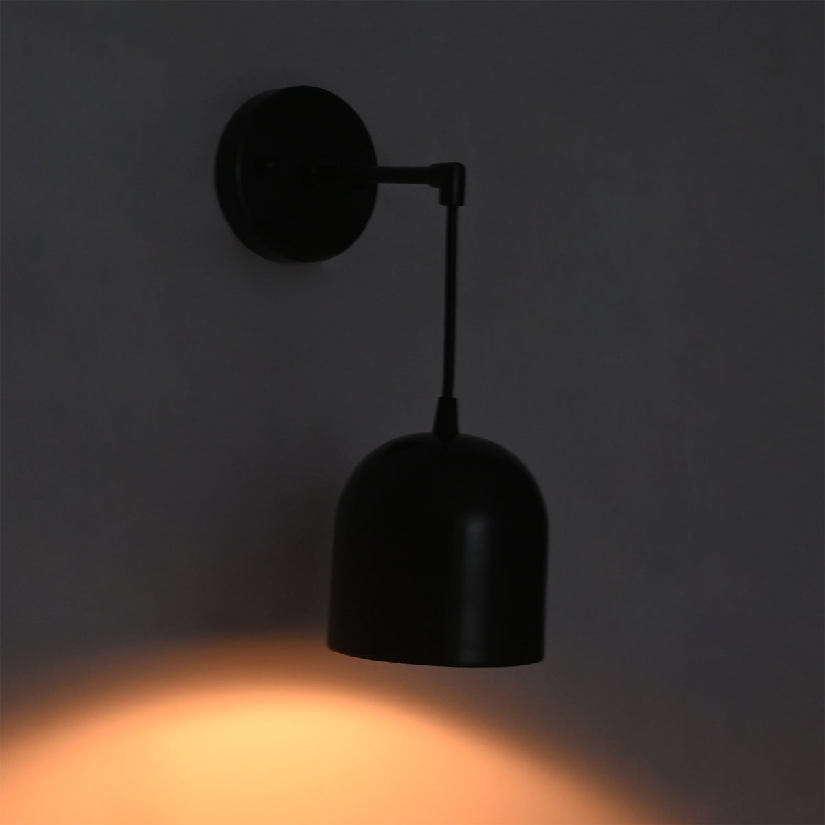 A modern black wall sconce with a dome-shaped shade and adjustable arm, mounted on a dark gray wall and casting a warm orange light downward.