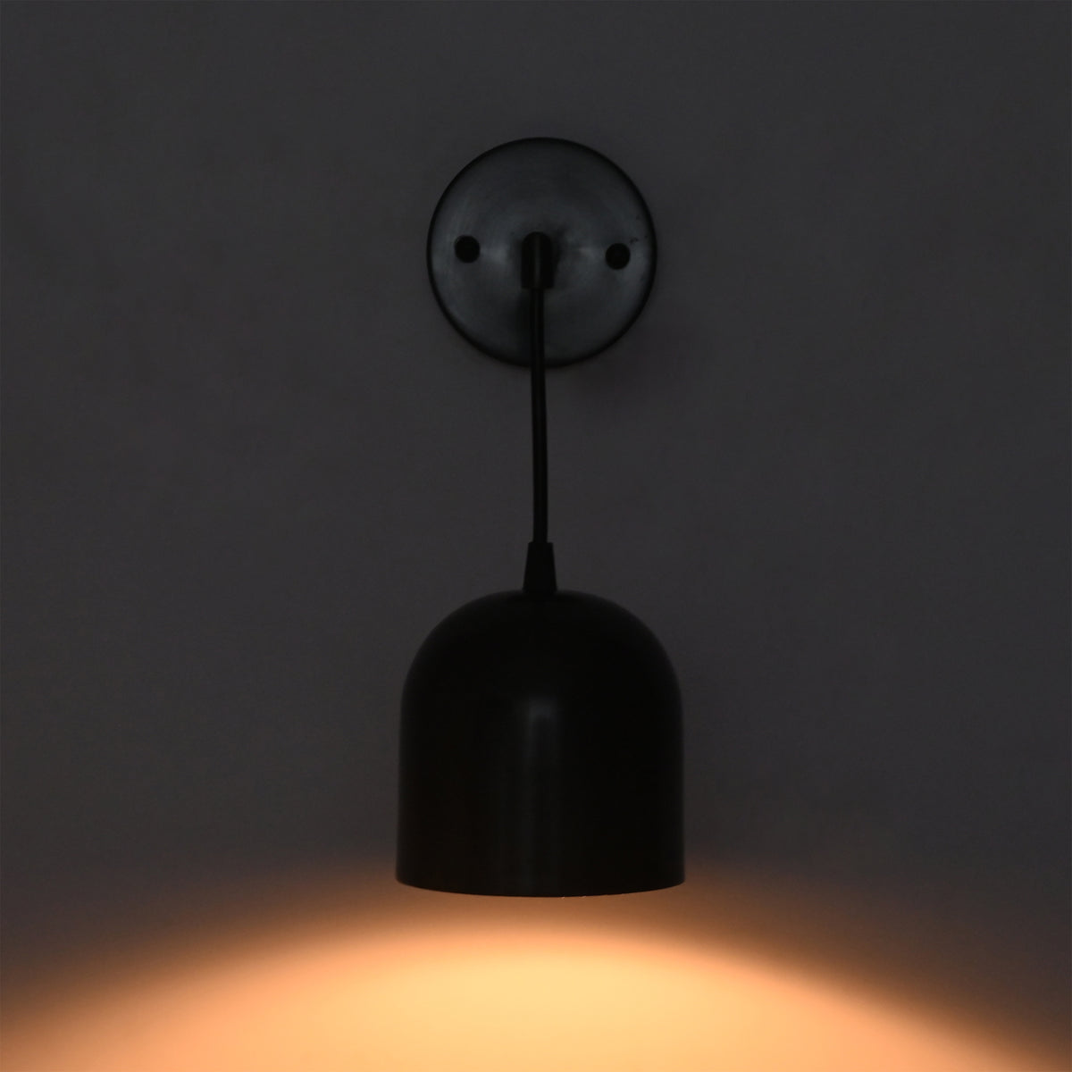 A modern black pendant wall light with a dome-shaped shade and circular wall plate, shown illuminated against a dark gray wall casting a warm orange glow.
