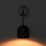 A modern black pendant wall light with a dome-shaped shade and circular wall plate, shown illuminated against a dark gray wall casting a warm orange glow.