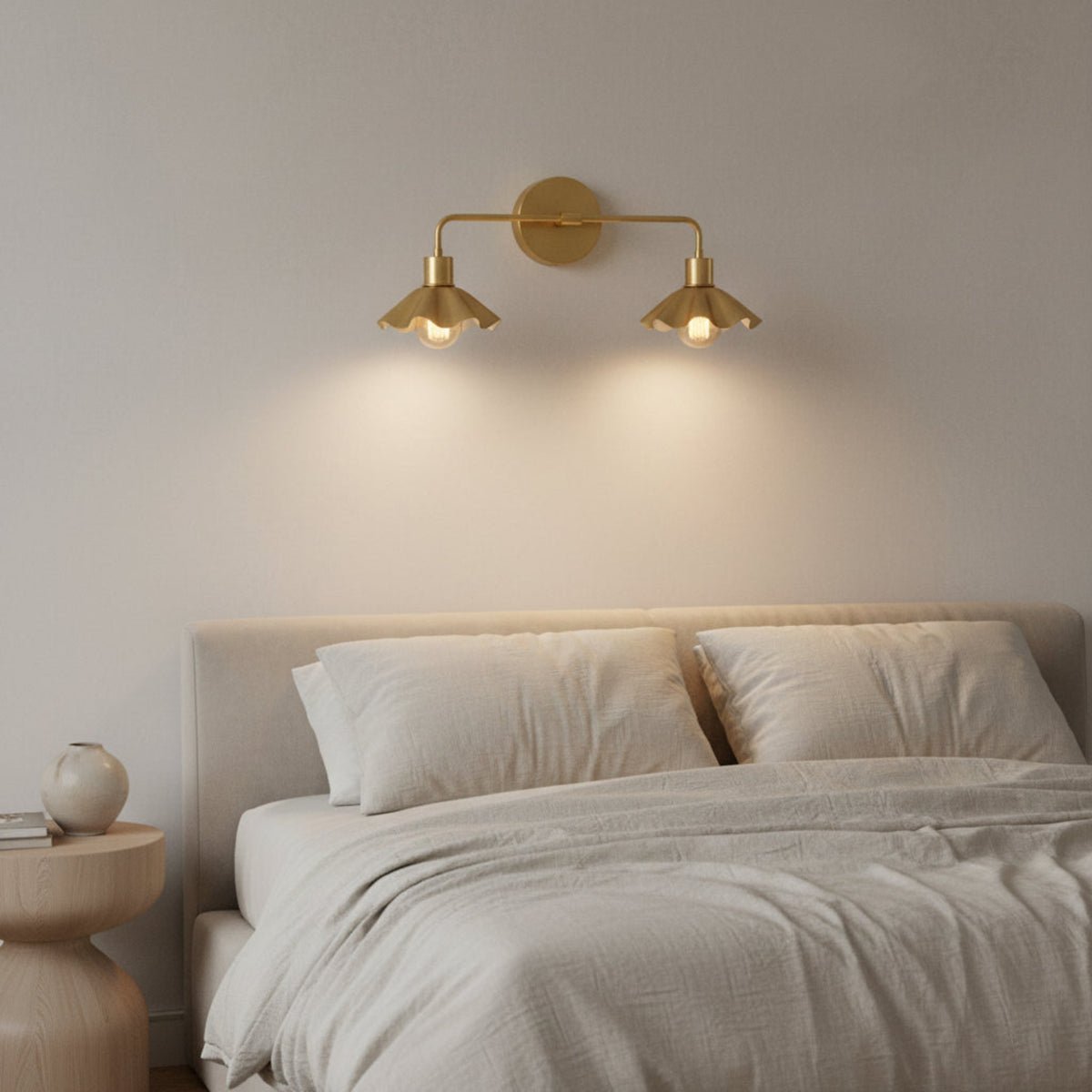 A modern two-light wall sconce in a brushed brass or gold finish, featuring a circular backplate, a horizontal arm, and two downward-facing flared shades with visible bulbs inside, shown against a plain white background.