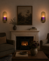A cozy living room scene with a fireplace, two upholstered armchairs, a coffee table, wall sconces with purple candlelight, and a framed landscape painting above the mantel.