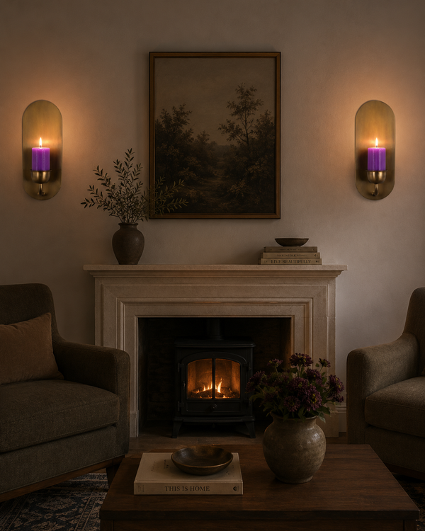 A cozy living room scene with a fireplace, two upholstered armchairs, a coffee table, wall sconces with purple candlelight, and a framed landscape painting above the mantel.