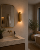 A modern bathroom interior featuring a warm brass wall sconce mounted on a textured beige plaster wall above a marble sink vanity with a gold-framed mirror.
