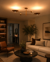 A modern brass three-arm ceiling light fixture with three bowl-shaped shades is illuminated in a warm, cozy living room setting with a beige sofa, brown leather chair, and dark marble coffee table.