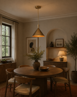 A warm, modern dining room featuring a round wooden table surrounded by six wooden chairs with light beige upholstery. A prominent brass-finished pendant light hangs over the table, casting a warm glow.