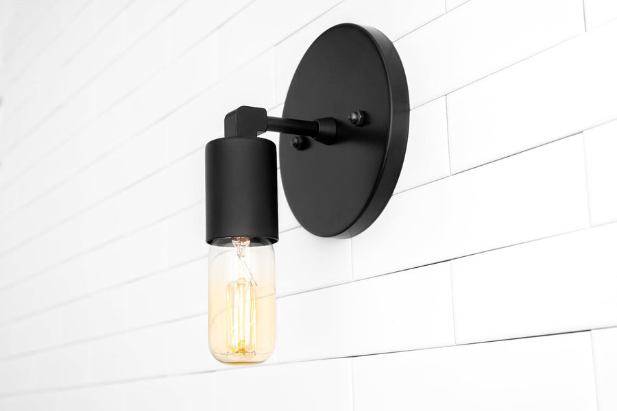 Matte black wall sconce with round backplate and capsule Edison bulb on white subway tile wall