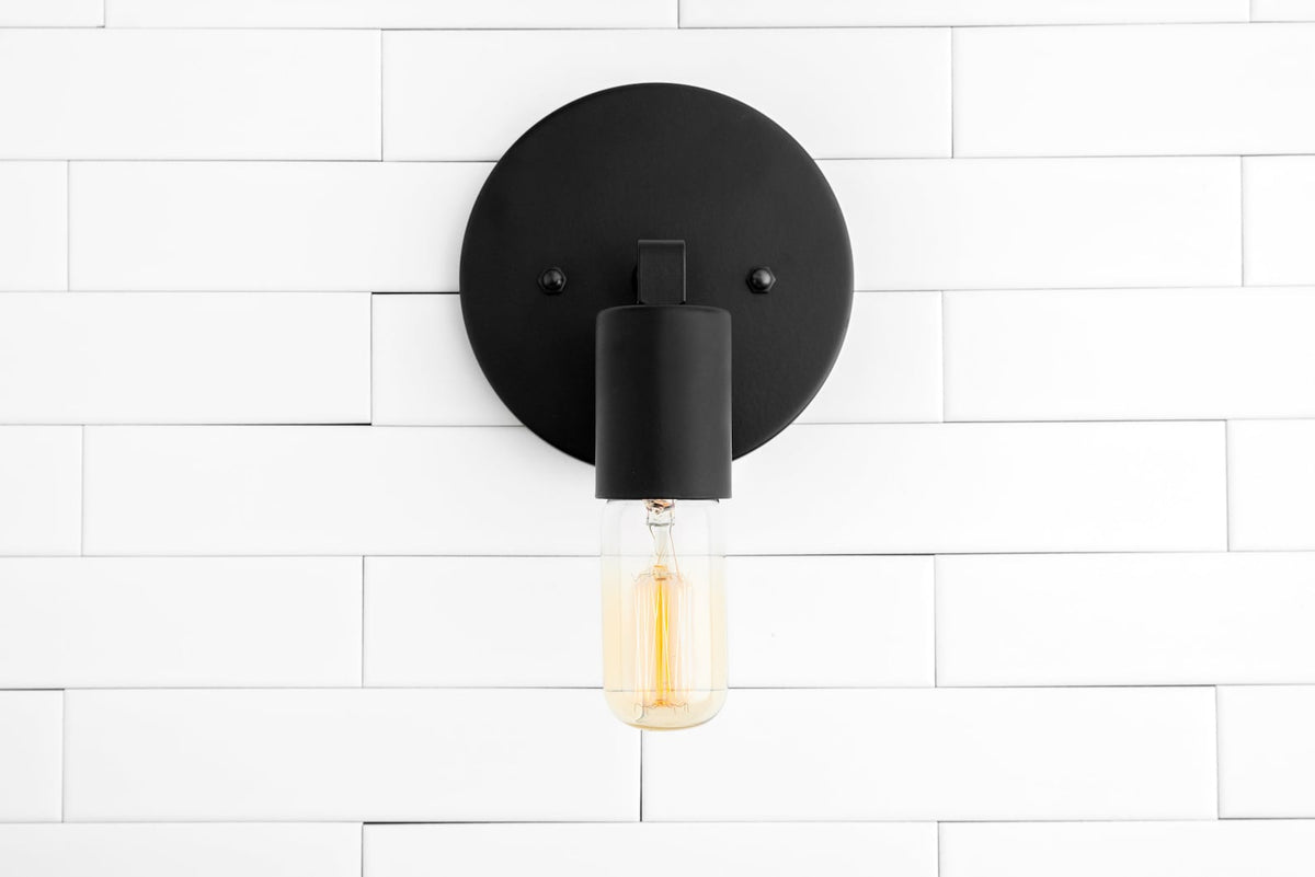 Minimal matte black wall sconce front view with round backplate and glowing capsule Edison bulb on white tile wall