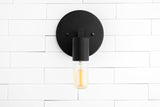 Minimal matte black wall sconce front view with round backplate and glowing capsule Edison bulb on white tile wall