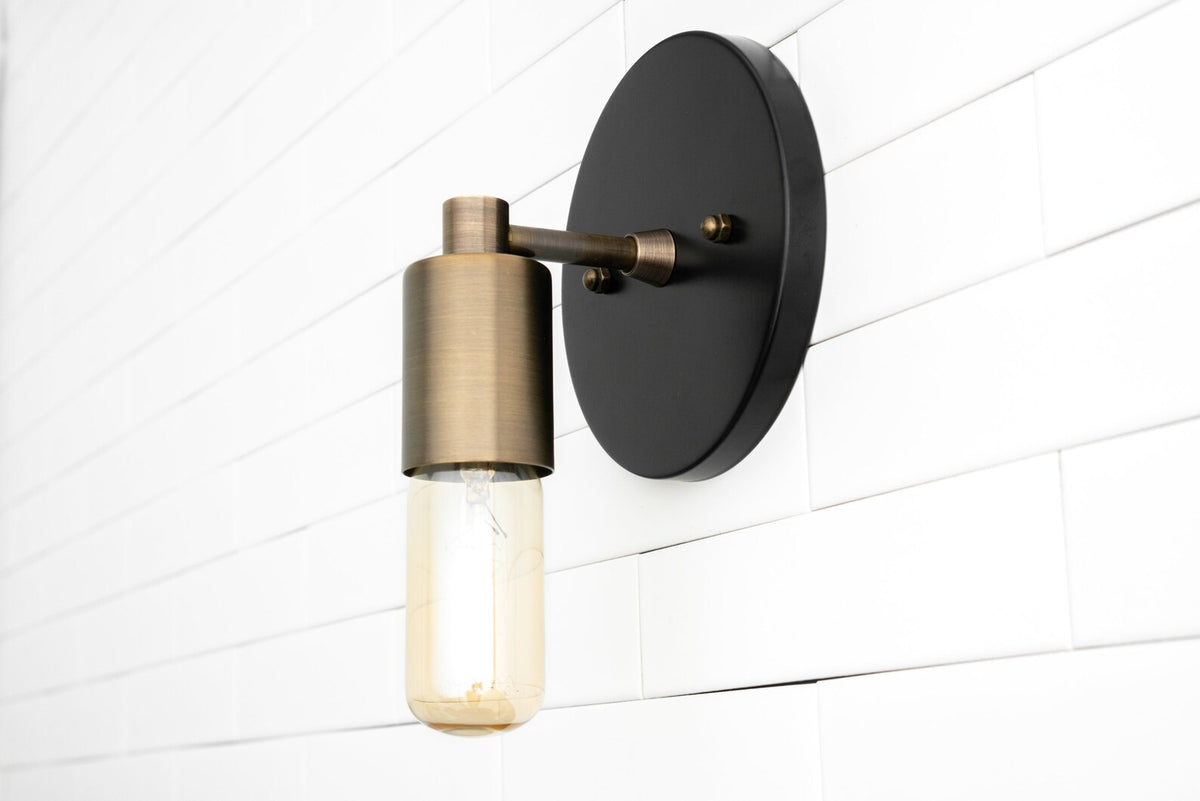 Modern wall sconce with matte black backplate and brushed brass cylinder socket holding capsule Edison bulb on white tile wall