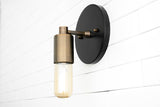 Modern wall sconce with matte black backplate and brushed brass cylinder socket holding capsule Edison bulb on white tile wall