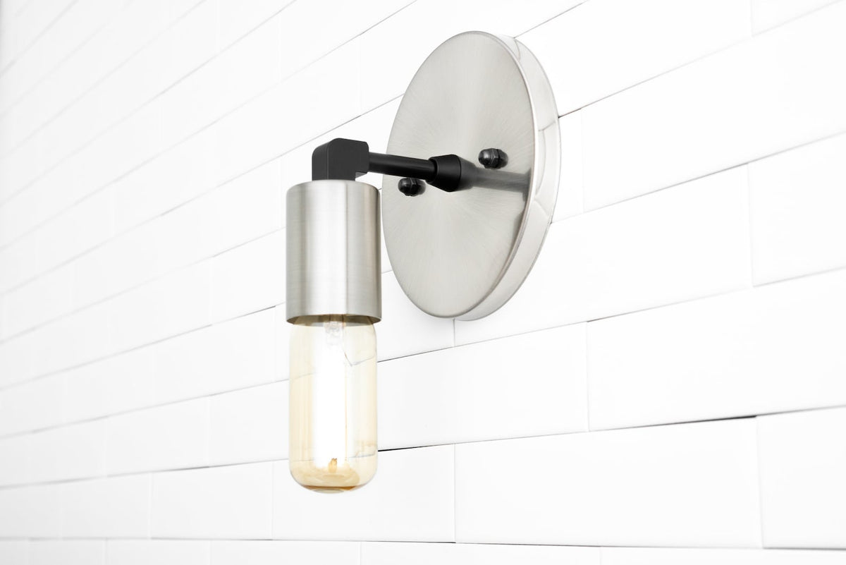 Brushed nickel wall sconce with round backplate and capsule Edison bulb, modern minimalist industrial style on white tile wall