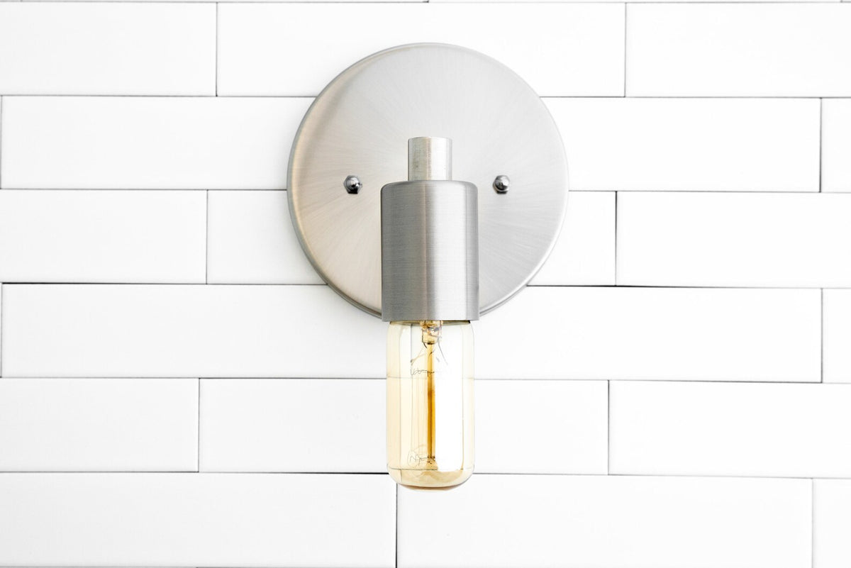Brushed nickel wall sconce front view with round backplate and capsule Edison bulb on white tiled wall