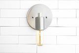 Brushed nickel wall sconce front view with round backplate and capsule Edison bulb on white tiled wall