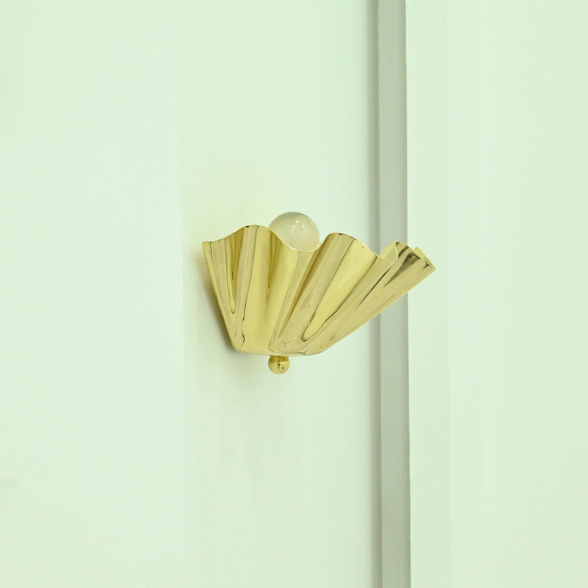  Brooklyn single-light polished brass wall sconce featuring an elegant folded fan-shaped metal shade mounted on a light-colored wall in natural daylight.