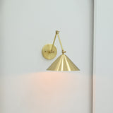 Adelaide 1-light wall sconce featuring a brushed brass cone shade, geometric articulated-style arms and circular wall plate against a white wall.