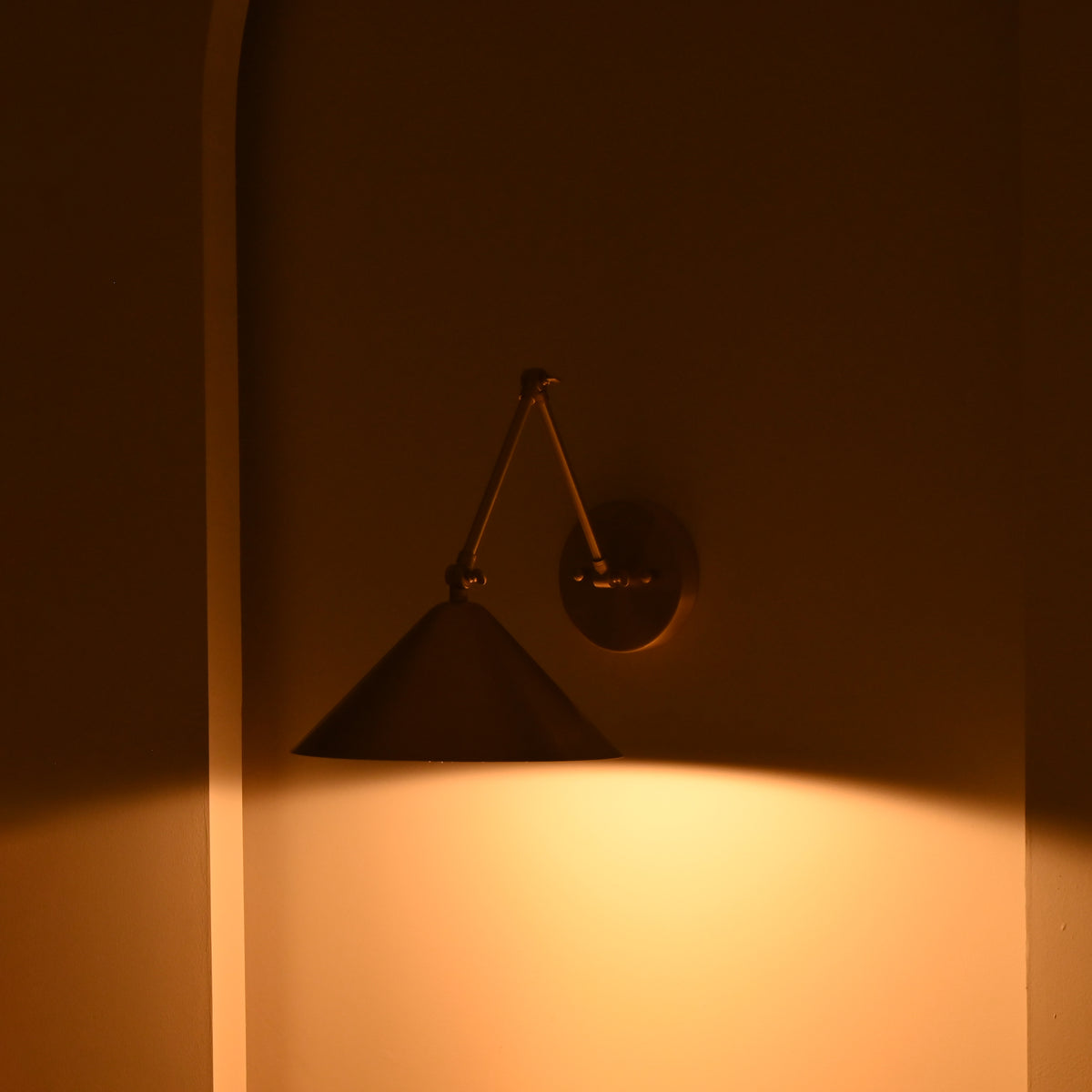 Adelaide brass cone wall sconce casting a focused pool of warm light downward in a dark, atmospheric interior with a sculptural silhouette.