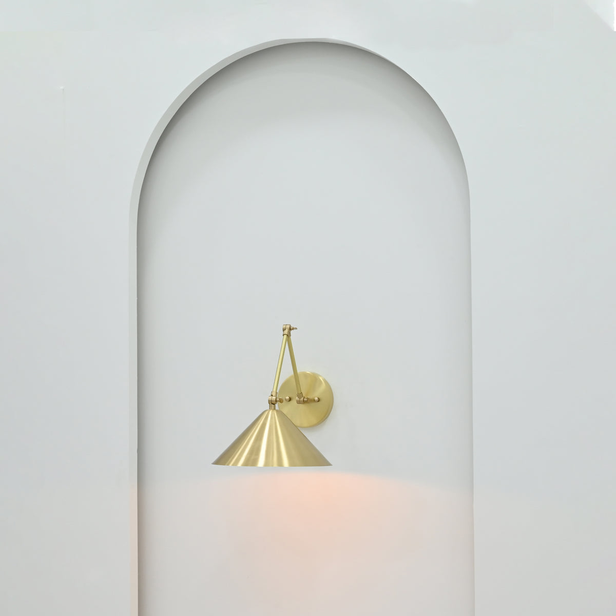 Adelaide brass cone wall sconce mounted inside a minimalist white arched wall niche with elegant round backplate and downward-facing metal shade.