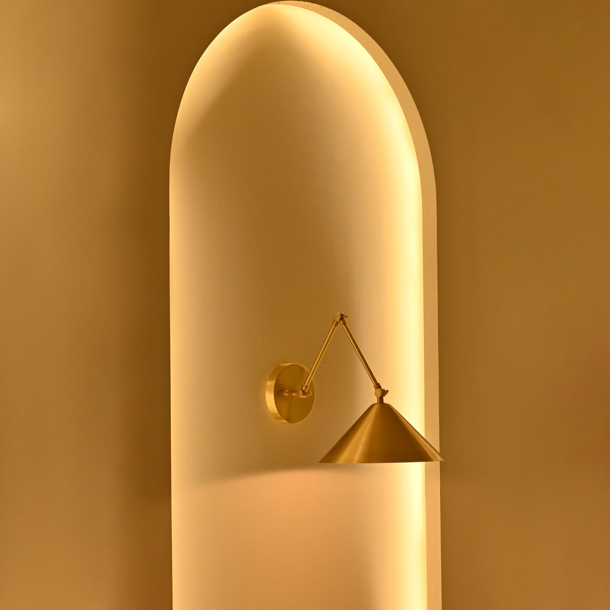Adelaide 1-light brass wall sconce with geometric double-arm construction and cone-shaped shade against a softly glowing golden arched wall.