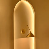 Adelaide 1-light brass wall sconce with geometric double-arm construction and cone-shaped shade against a softly glowing golden arched wall.