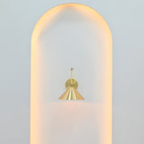 Adelaide brass cone wall light illuminating a minimalist arched wall niche with soft warm lighting and a sculptural metal shade.