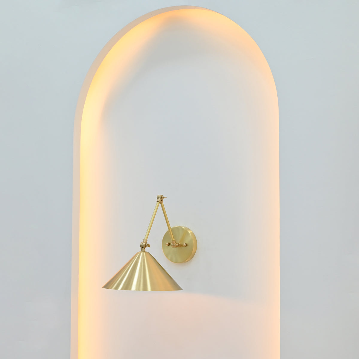 Adelaide 1-light brass wall sconce with wide conical shade and angular metal arms creating soft downward illumination inside a white arched niche.