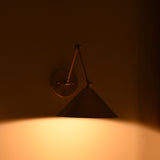 Adelaide 1-light brass cone wall sconce with angular arms and round mounting plate creating warm downward illumination against a dark wall.