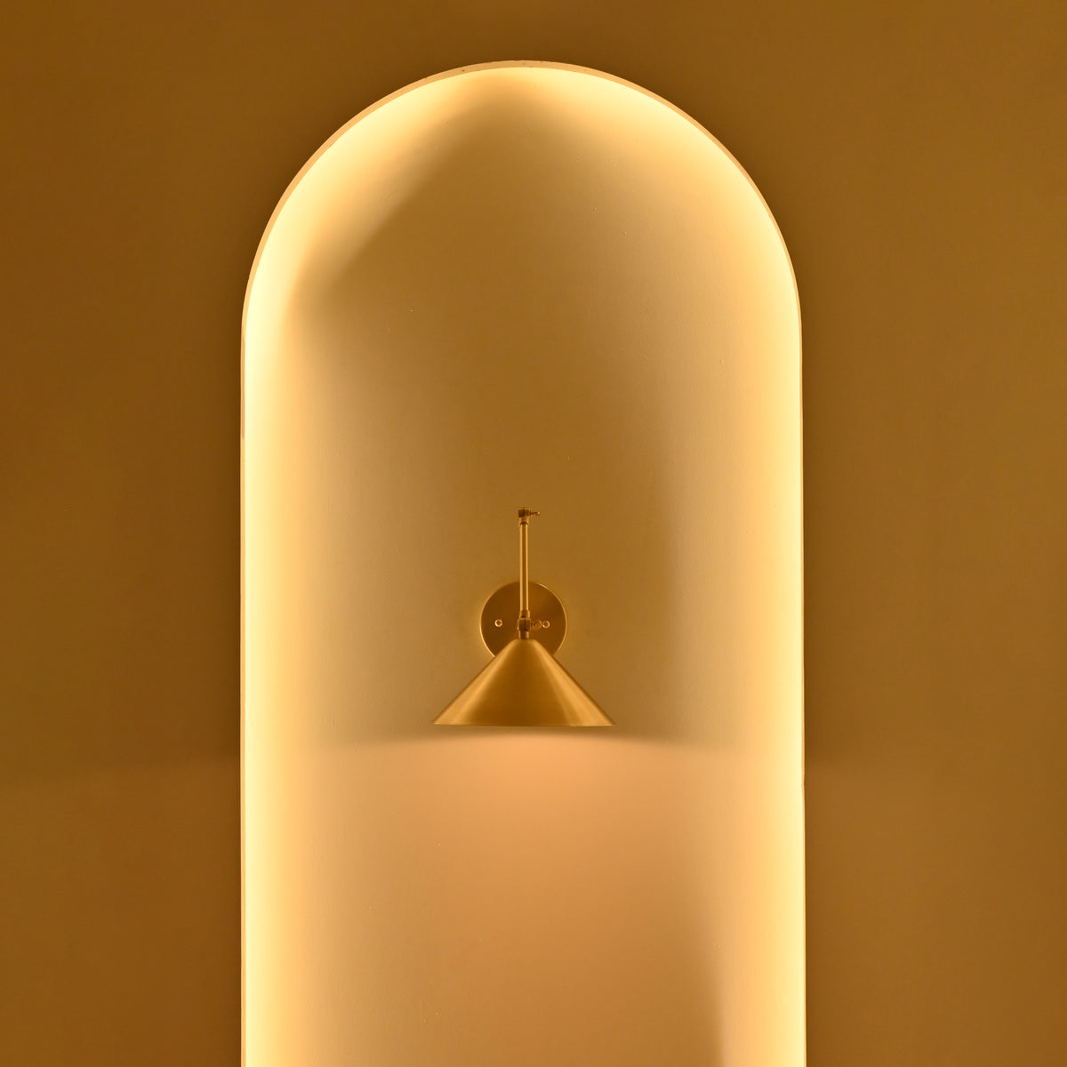 Adelaide brass cone wall sconce displayed in a warmly illuminated architectural niche with golden ambient lighting and elegant minimalist styling.