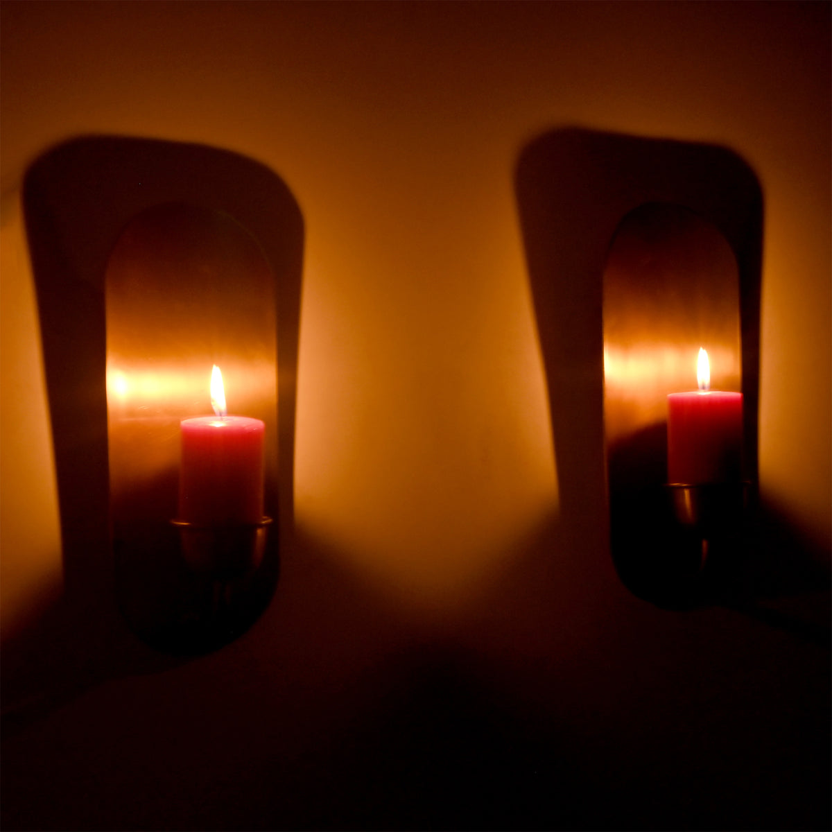 Pair of Alden modern brass wall-mounted candle sconces with lit pillar candles creating warm ambient lighting and soft shadows on a decorative interior wall.