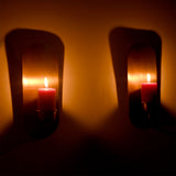 Pair of Alden modern brass wall-mounted candle sconces with lit pillar candles creating warm ambient lighting and soft shadows on a decorative interior wall.