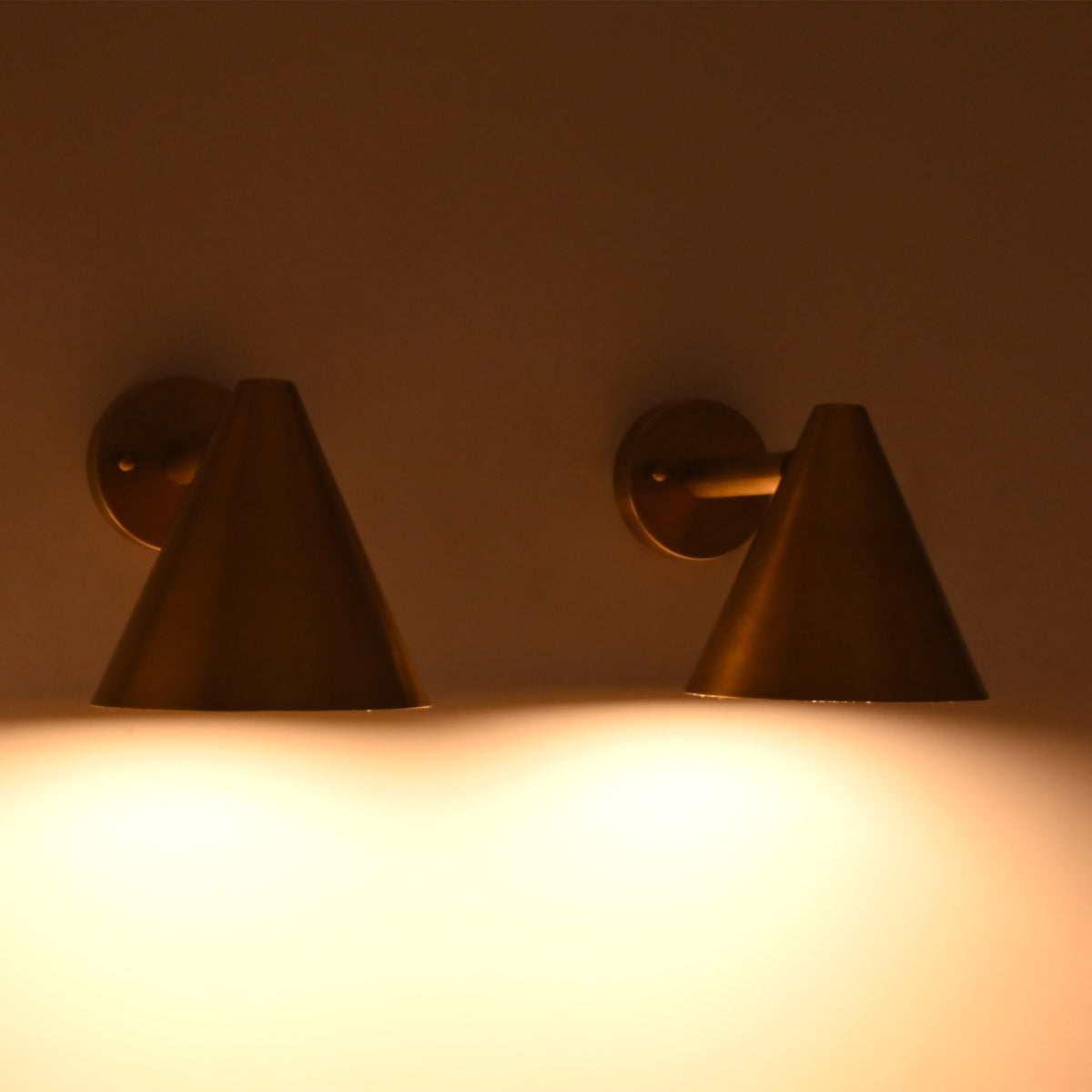 Front view of the Aria modern tapered cone brass wall sconce illuminated, casting a warm focused downward glow.