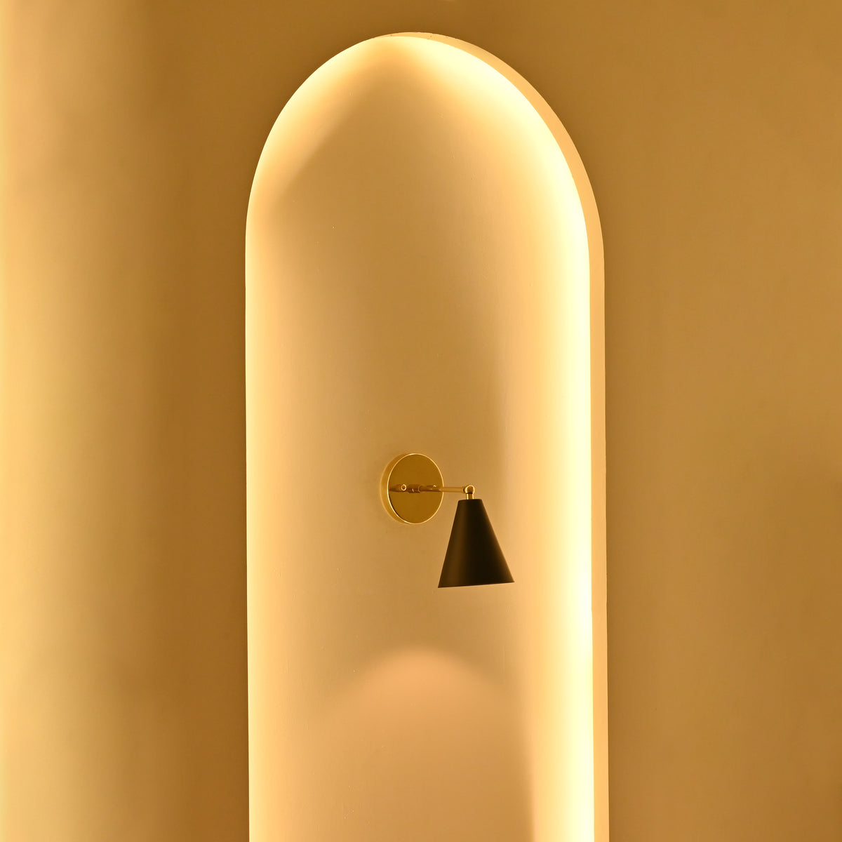 Aspen single-light black cone wall sconce with brass mounting plate displayed in a softly illuminated architectural niche during evening lighting.