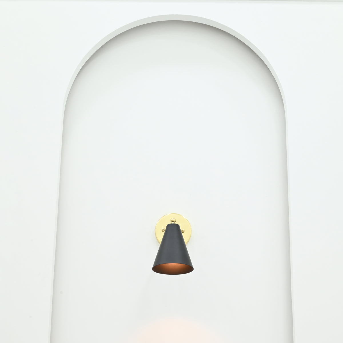 Front view of Aspen single-light black cone wall sconce with circular polished brass backplate installed inside a minimalist white arched wall niche.