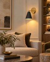 Aspen black and gold cone wall sconce providing warm accent lighting above a neutral sofa in a contemporary living room with wooden shelving and elegant decor.