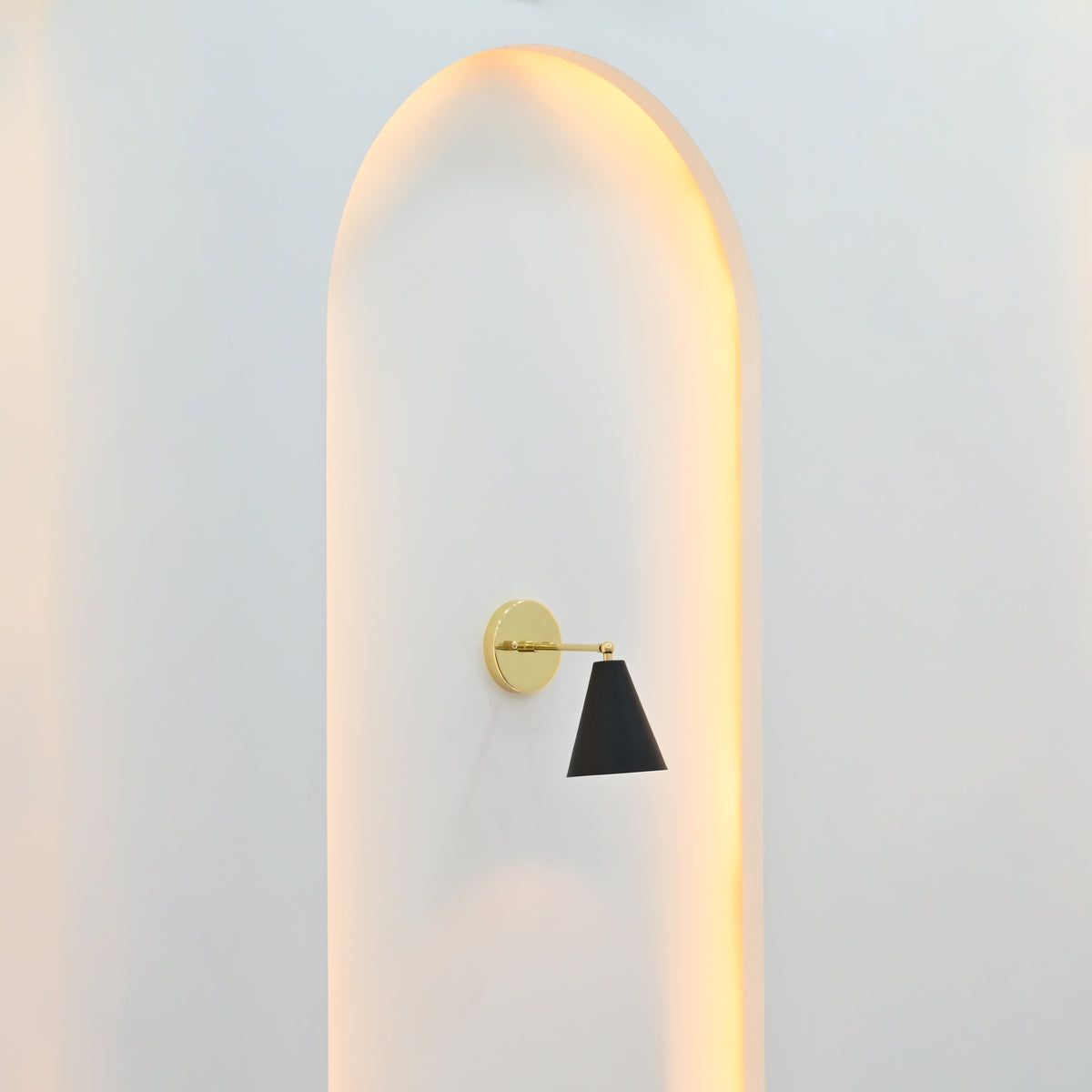  Angled view of Aspen matte black cone wall light with polished brass arm and round backplate displayed against a softly illuminated arched wall.