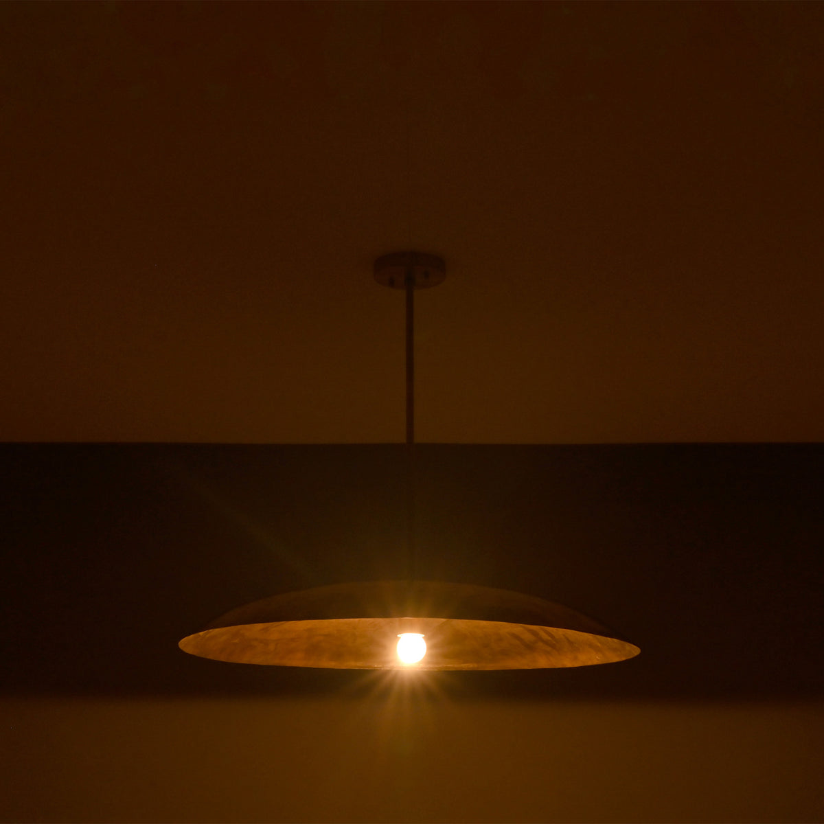 Wide saucer pendant light displayed in low light with warm exposed bulb and dramatic glow.