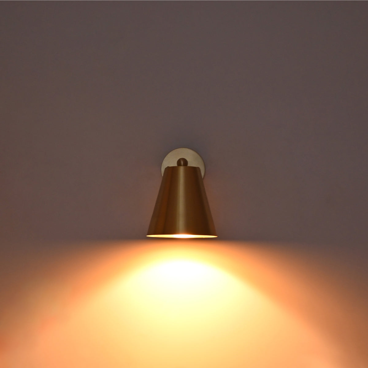 Front view of the Aurora satin brass tapered wall sconce illuminated with a warm downward glow creating soft ambient wall lighting.