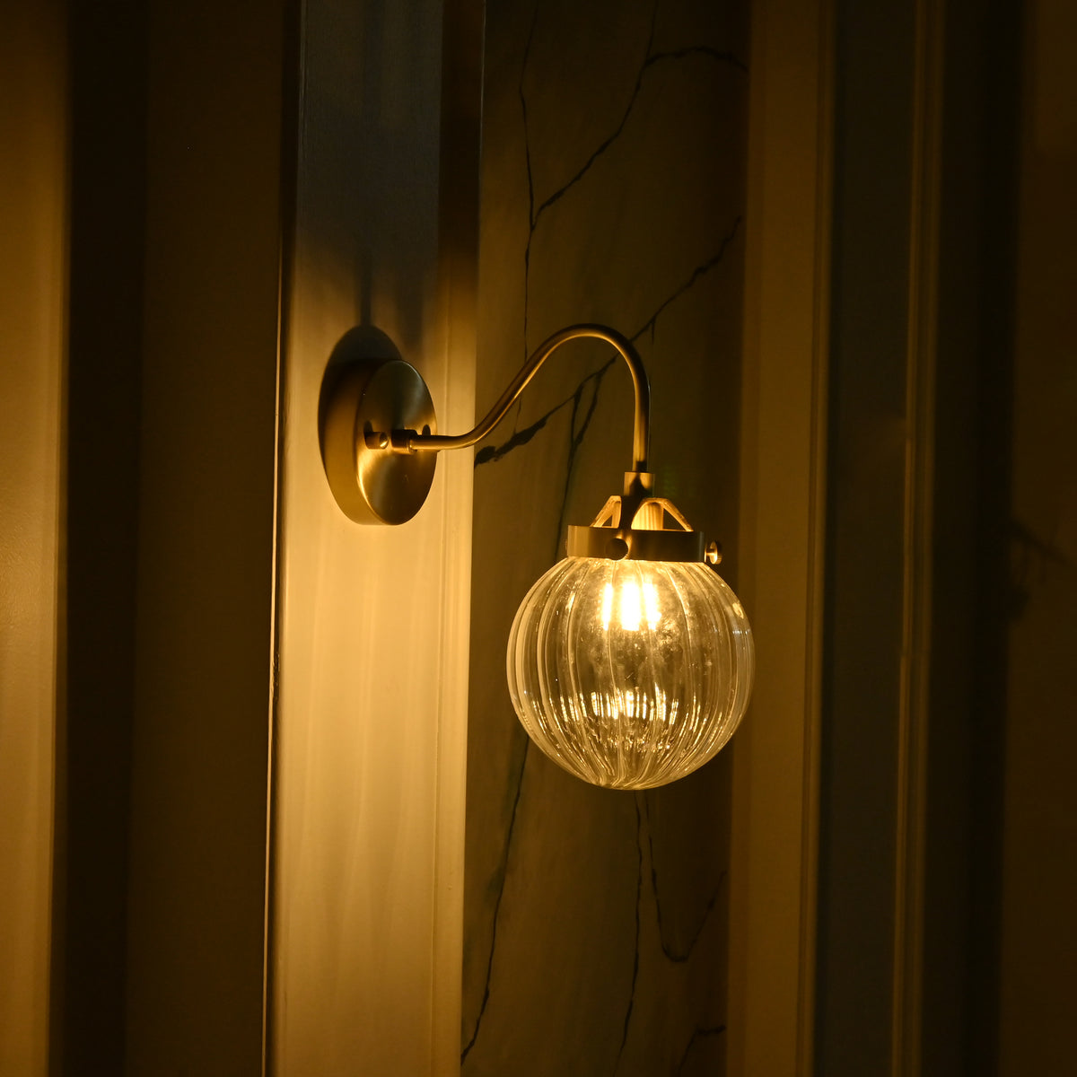 Side profile of Austin curved brass wall sconce with a clear textured glass globe illuminated in warm golden light against a dark wall.