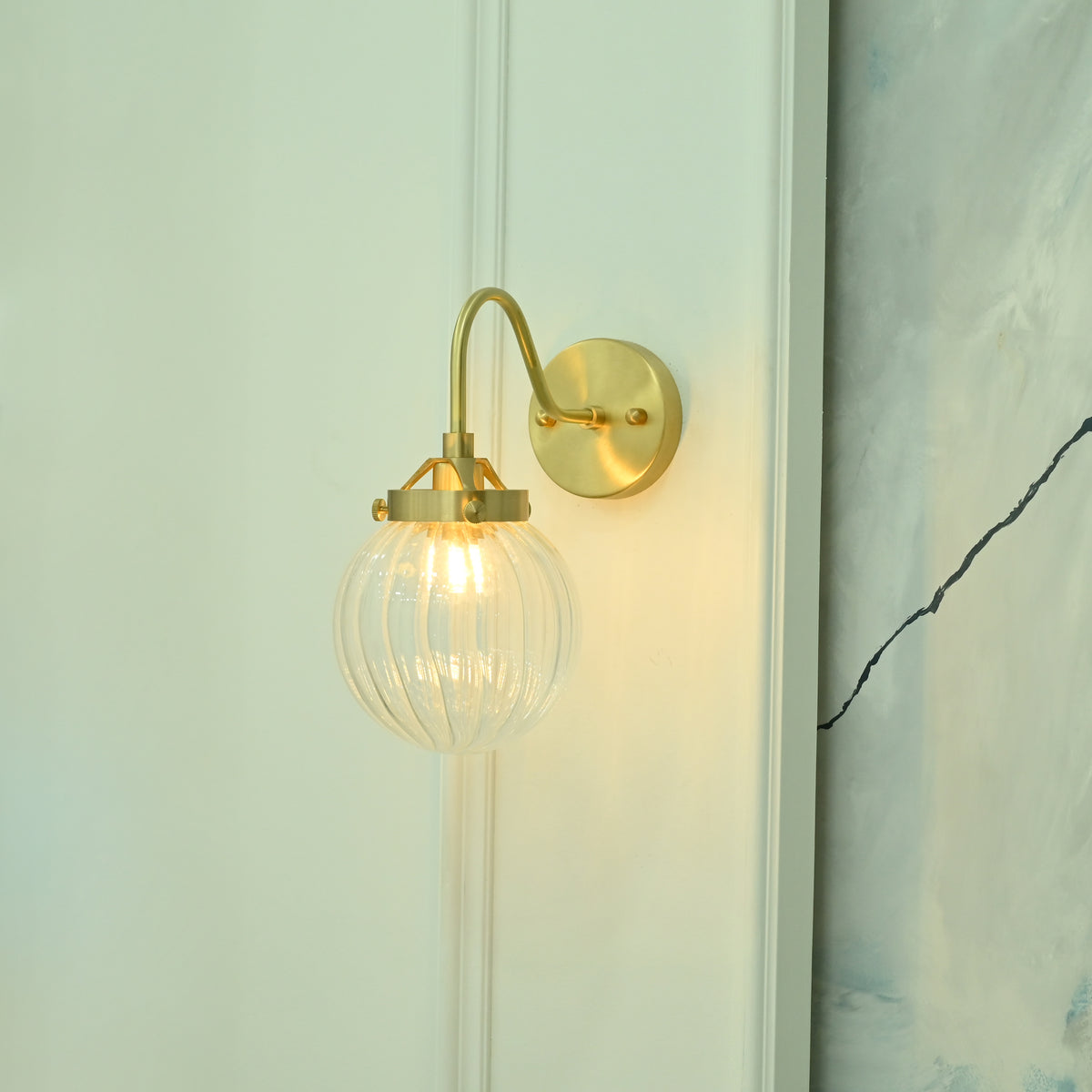 Angled view of Austin single-light wall sconce with an elegant curved brass arm, round wall backplate, and transparent ribbed glass globe.