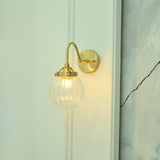 Angled view of Austin single-light wall sconce with an elegant curved brass arm, round wall backplate, and transparent ribbed glass globe.